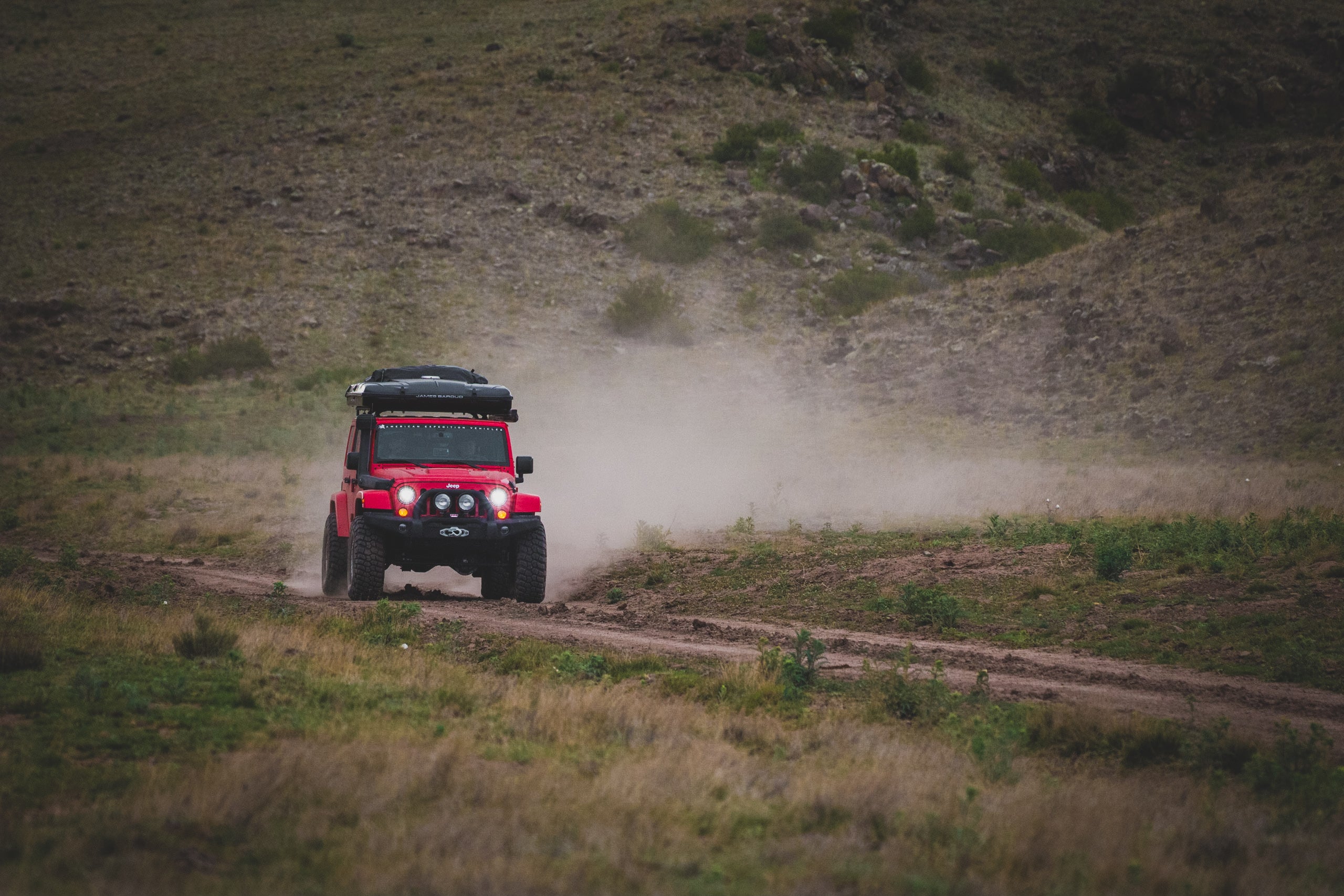 Red off-road vehicle driving on a dirt path in a grassy, hilly landscape